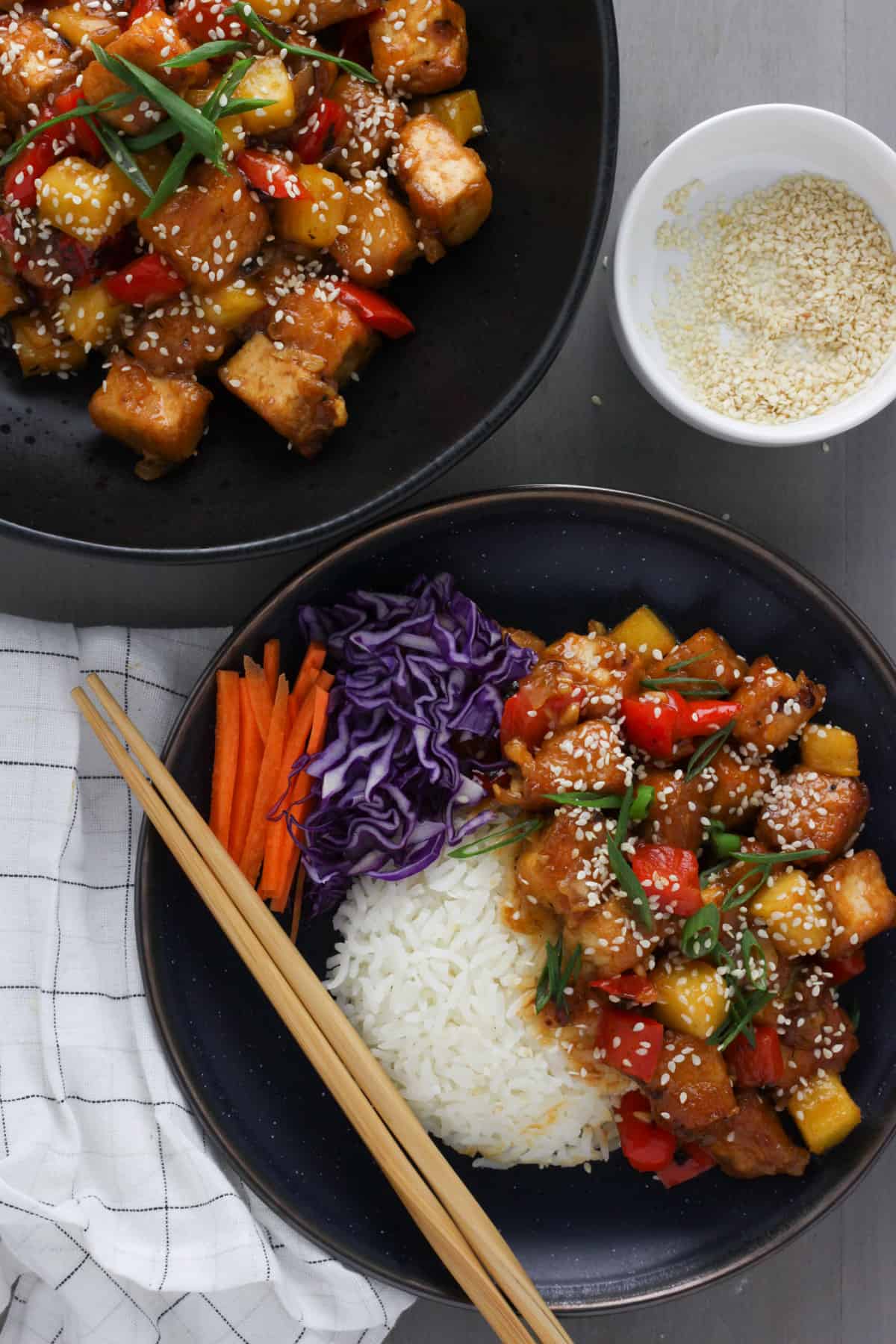 A dark blue bowl with white rice, thinly sliced red cabbage and matchsticks of carrots with cubed tofu, red pepper and onions in a sweet and sour sauce, all topped with sesame seeds and scallions. There are bamboo chopsticks on the edge of the bowl and another platter of sweet and sour tofu off center, along with a small bowl of sesame seeds.