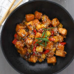 A black bowl with sweet and sour tofu topped with sesame seeds and scallions and a small white bowl with sesame seeds and a napkin just off center.