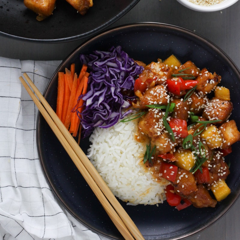 A black bowl with white rice topped with sweet and sour tofu, sesame seeds, cabbage and carrots.