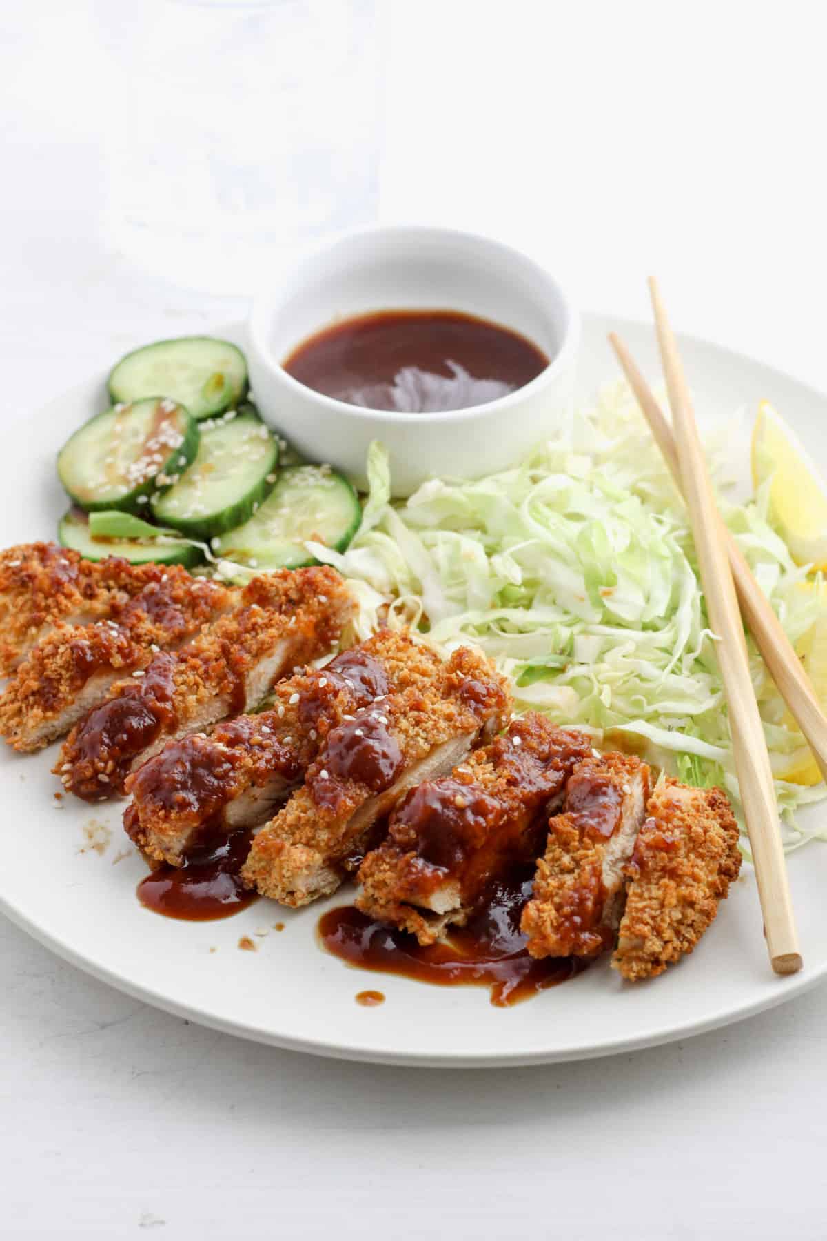 Air Fryer Chicken Katsu on a plate with shredded green cabbage, sliced cucumbers, and tonkatsu sauce in a small bowl with chopsticks on the side of the plate.