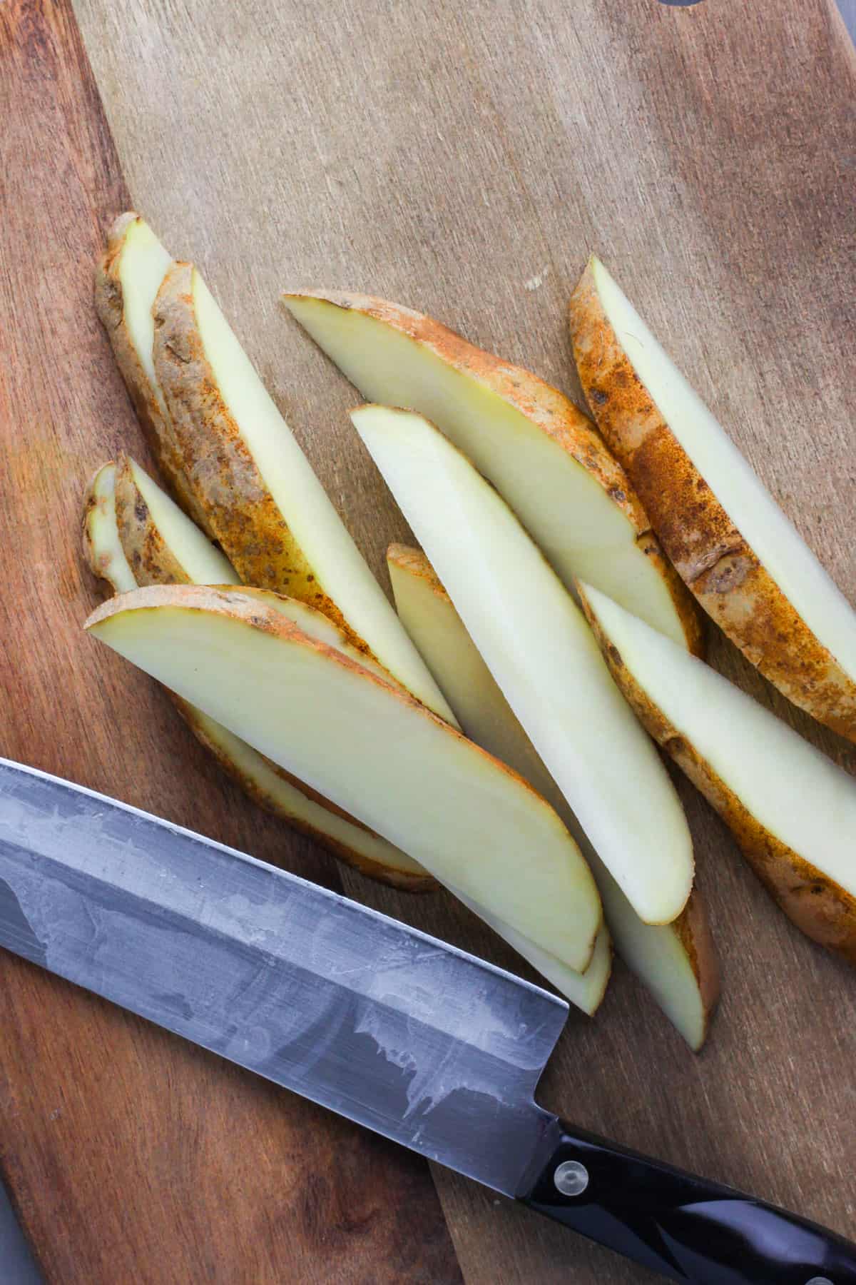 A cutting board with chef knife and russet potatoes cut into wedges.
