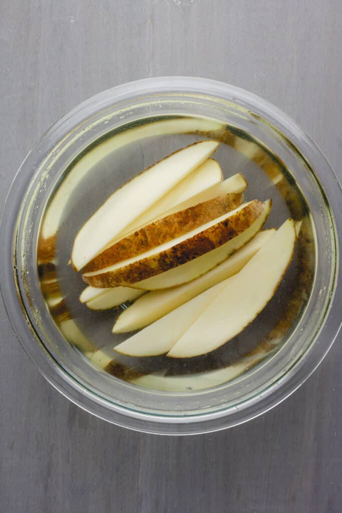 A glass mixing bowl with water cover wedges of russet potatoes.