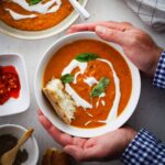 A bowl of lentil and red pepper soup held by two hands with another bowl of soup off center. The soup is topped with yogurt, basil and bread.