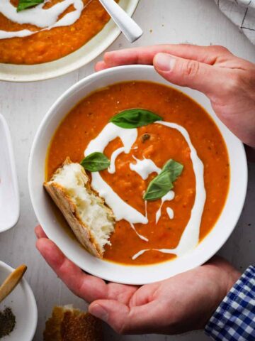 A bowl of lentil and red pepper soup held by two hands with another bowl of soup off center. The soup is topped with yogurt, basil and bread.