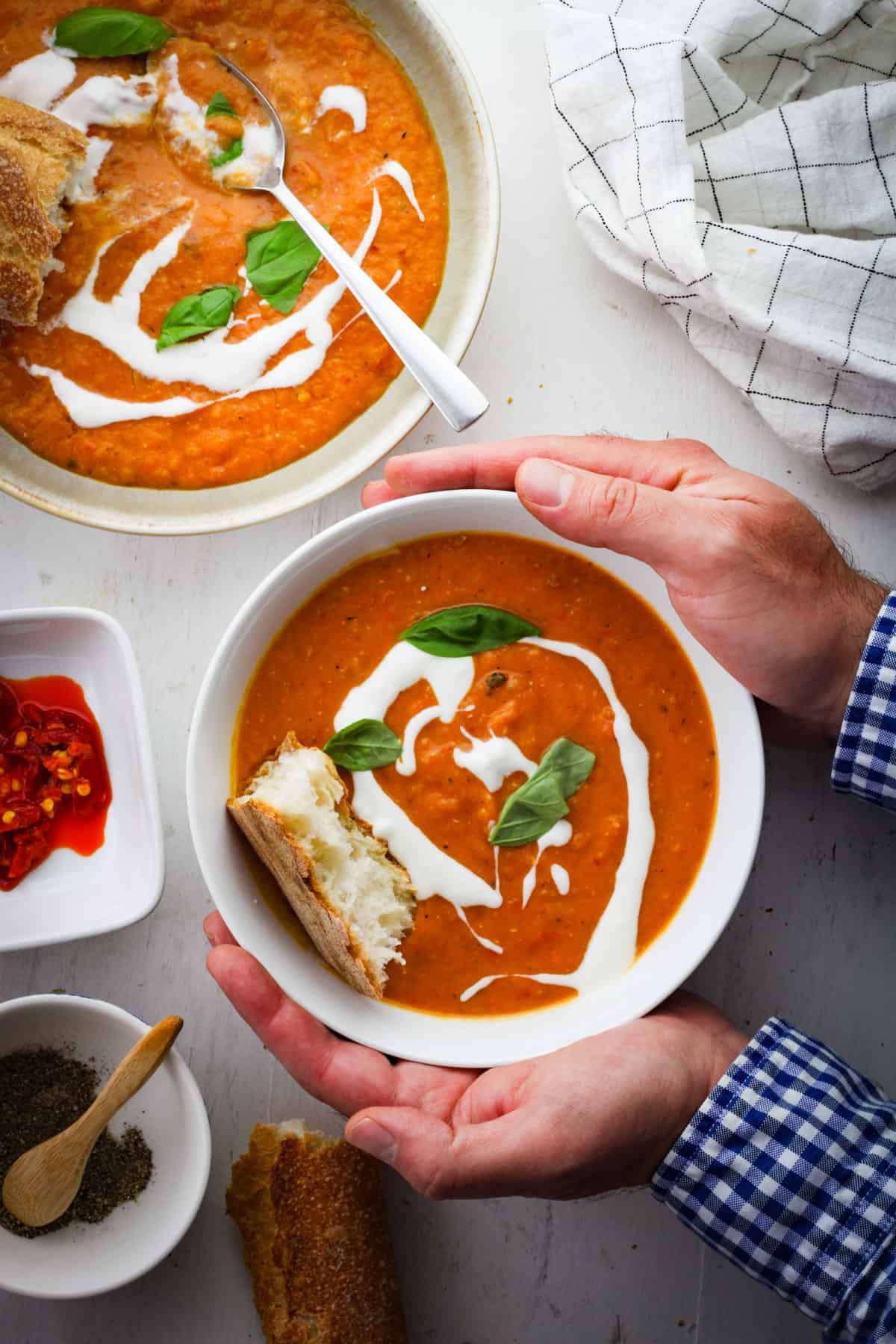 A bowl of lentil and red pepper soup held by two hands with another bowl of soup off center. The soup is topped with yogurt, basil and bread.