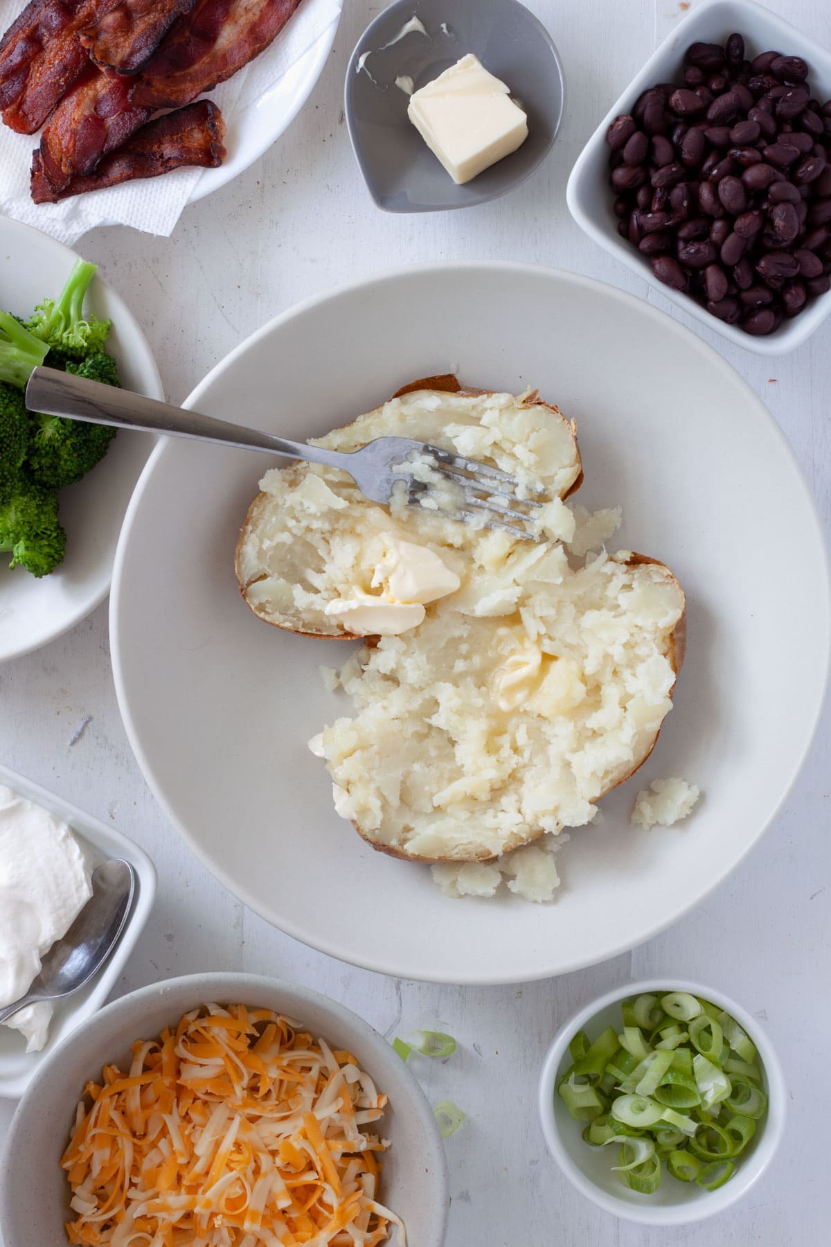 Butter mashed into a baked potato with ingredients for a loaded potato bowl along the edges.