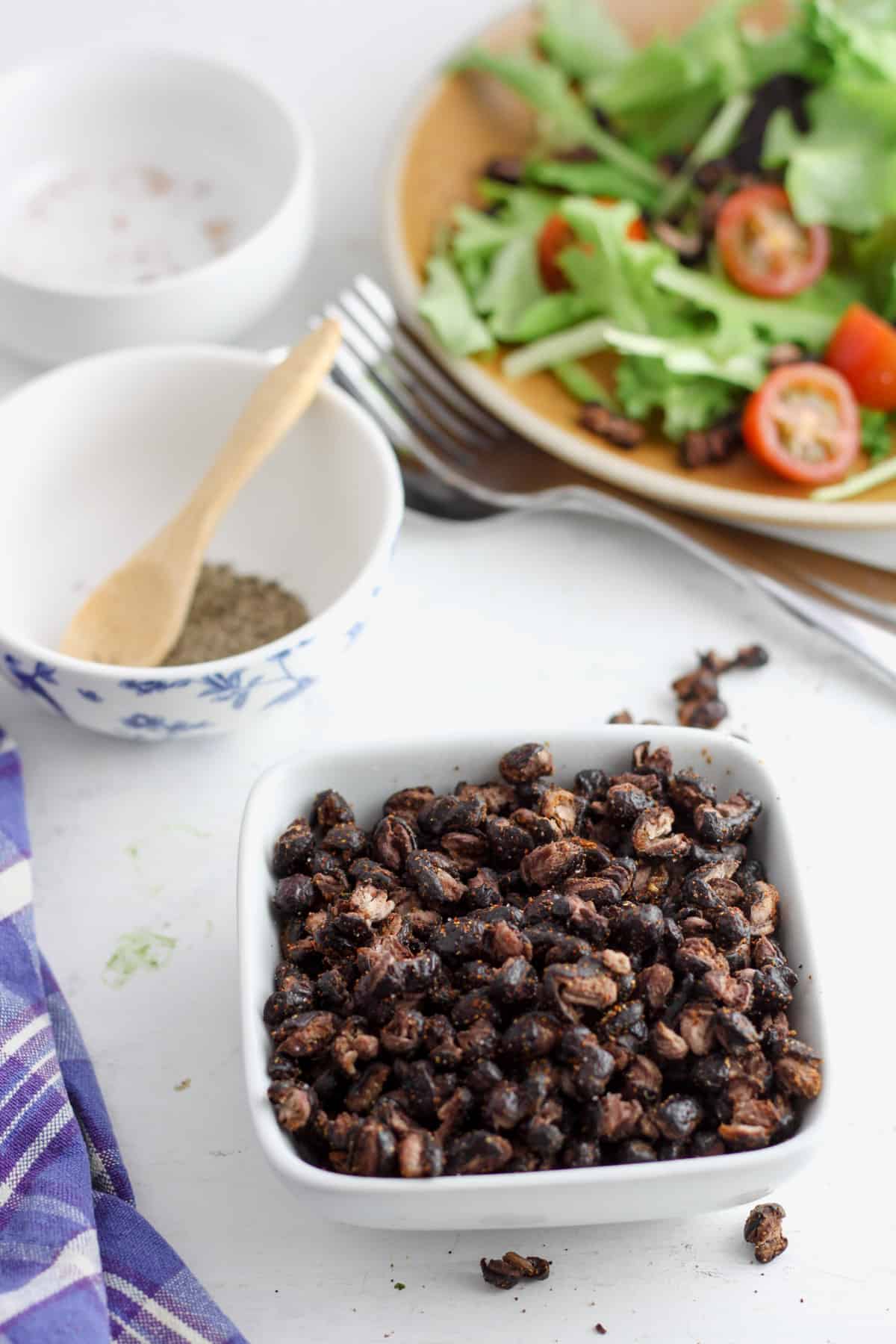 A small white bowl of roasted black beans with a salad off center and out of focus in the background.
