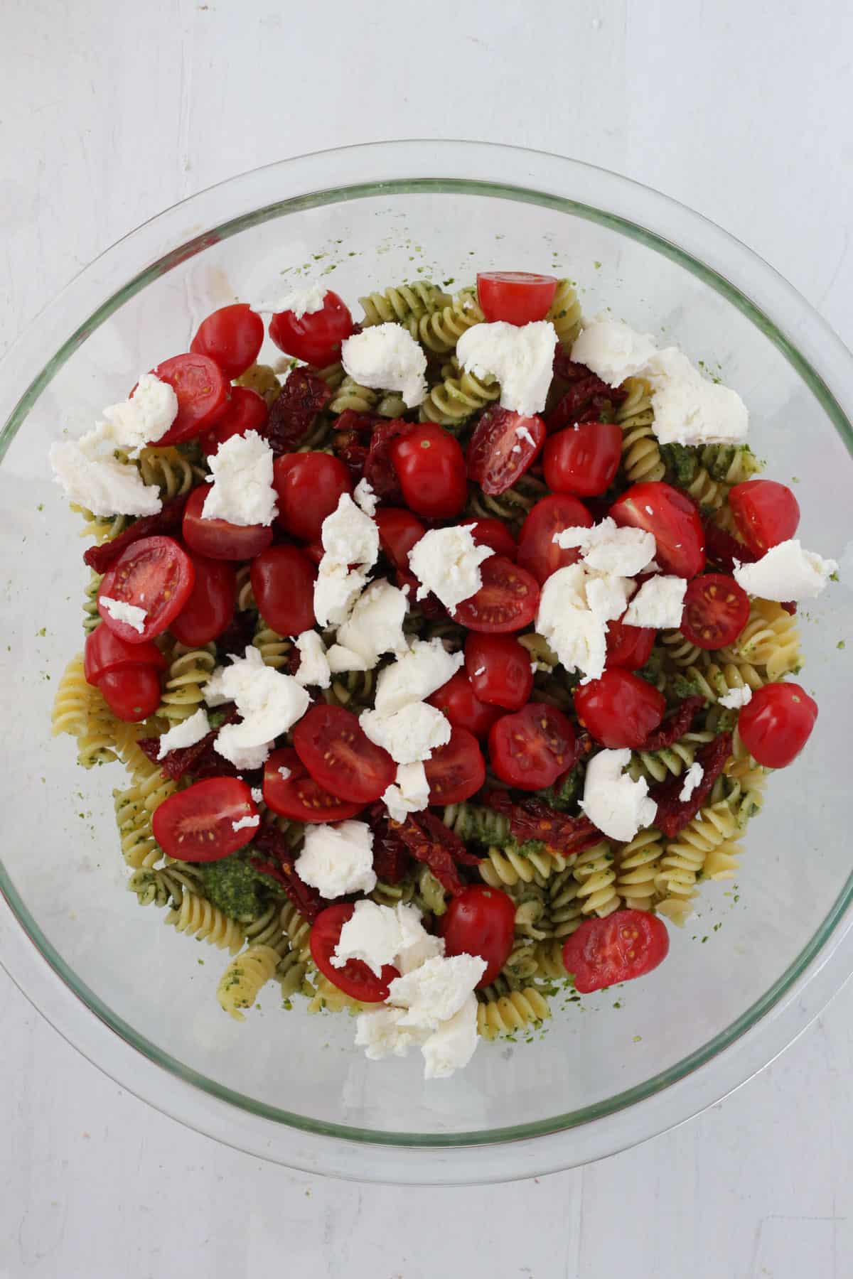 A large mixing bowl with pasta, pesto, sundried tomatoes, torn fresh mozzarella and halved cherry tomatoes.