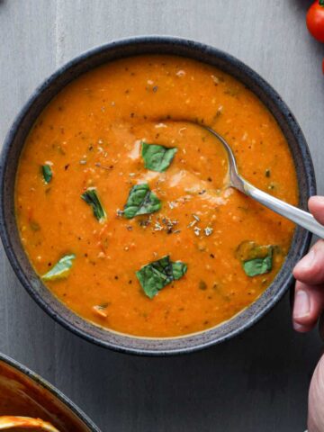 Cherry tomato soup in a gray bowl with a hand taking a spoonful.