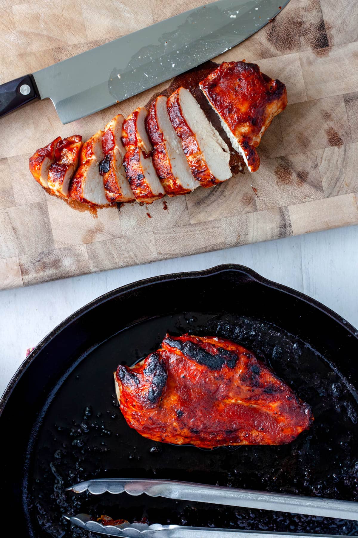 A bbq chicken breast getting sliced on a cutting board with another still in a cast iron skillet 