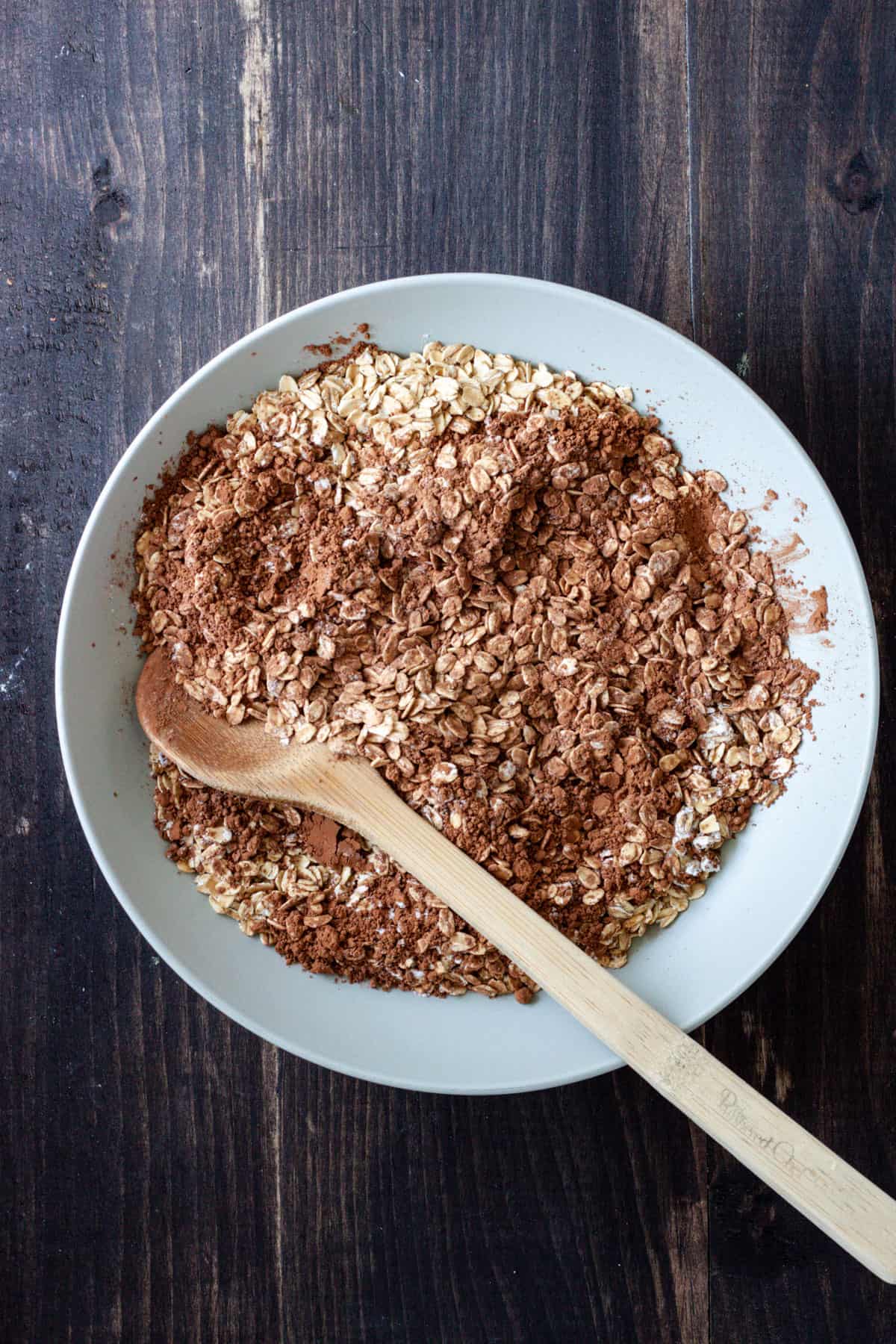 Oats and cocoa powder in a mixing bowl with a wooden spoon getting ready to mix it all together.