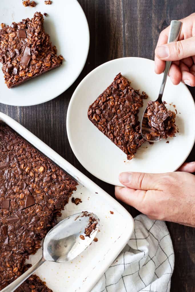 Chocolate Baked Oatmeal in a baking dish with two portions on small plates and a hand taking a spoonful from one portion.