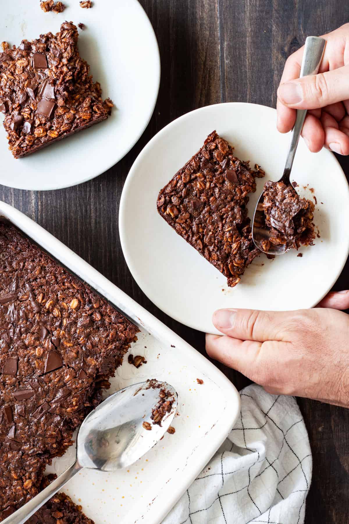 Chocolate Baked Oatmeal in a baking dish with two portions on small plates and a hand taking a spoonful from one portion.