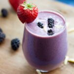 A glass of blackberry strawberry banana smoothie with a strawberry on the rim of the glass.