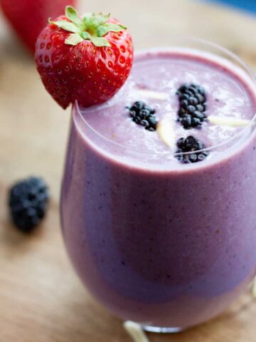 A glass of blackberry strawberry banana smoothie with a strawberry on the rim of the glass.