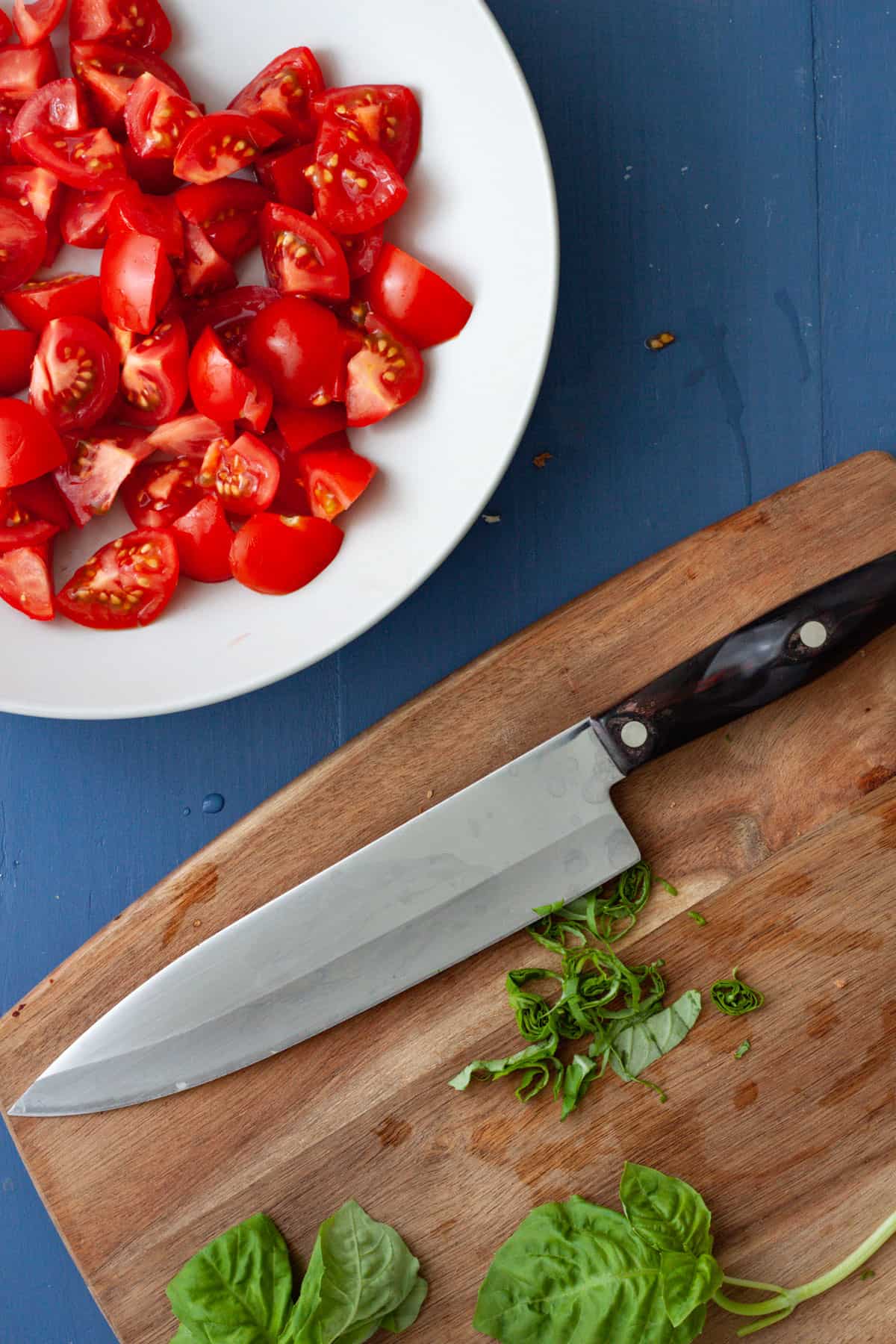 Basil getting sliced on a cutting board with a bowl of chopped tomatoes off center.