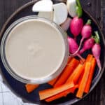 Miso and tahini dressing in a small bowl on a plate with a colorful assortment of vegetables.