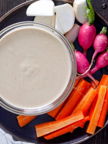 Miso and tahini dressing in a small bowl on a plate with a colorful assortment of vegetables.