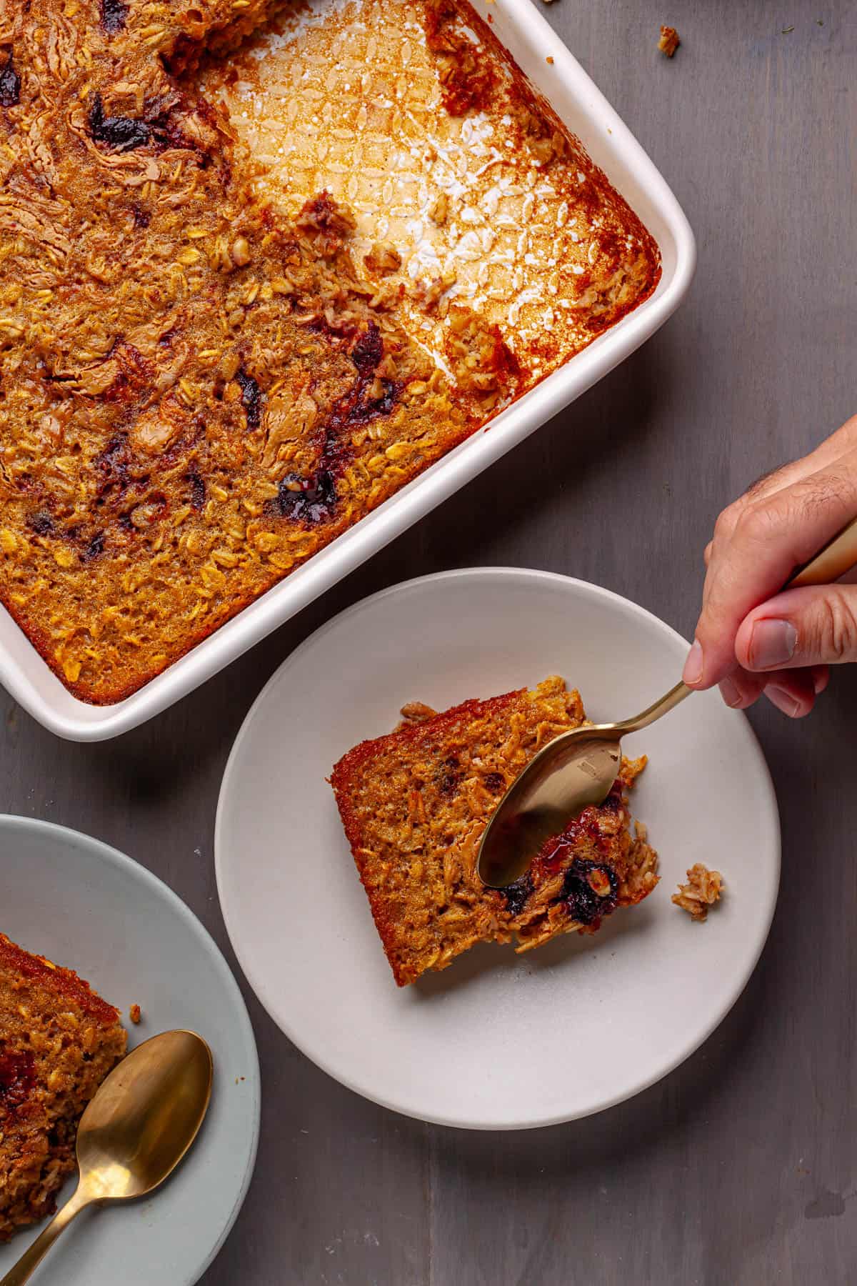 Peanut butter and jam baked oatmeal in a baking dish with two servings on small plates and a spoon taking a bit off one serving.