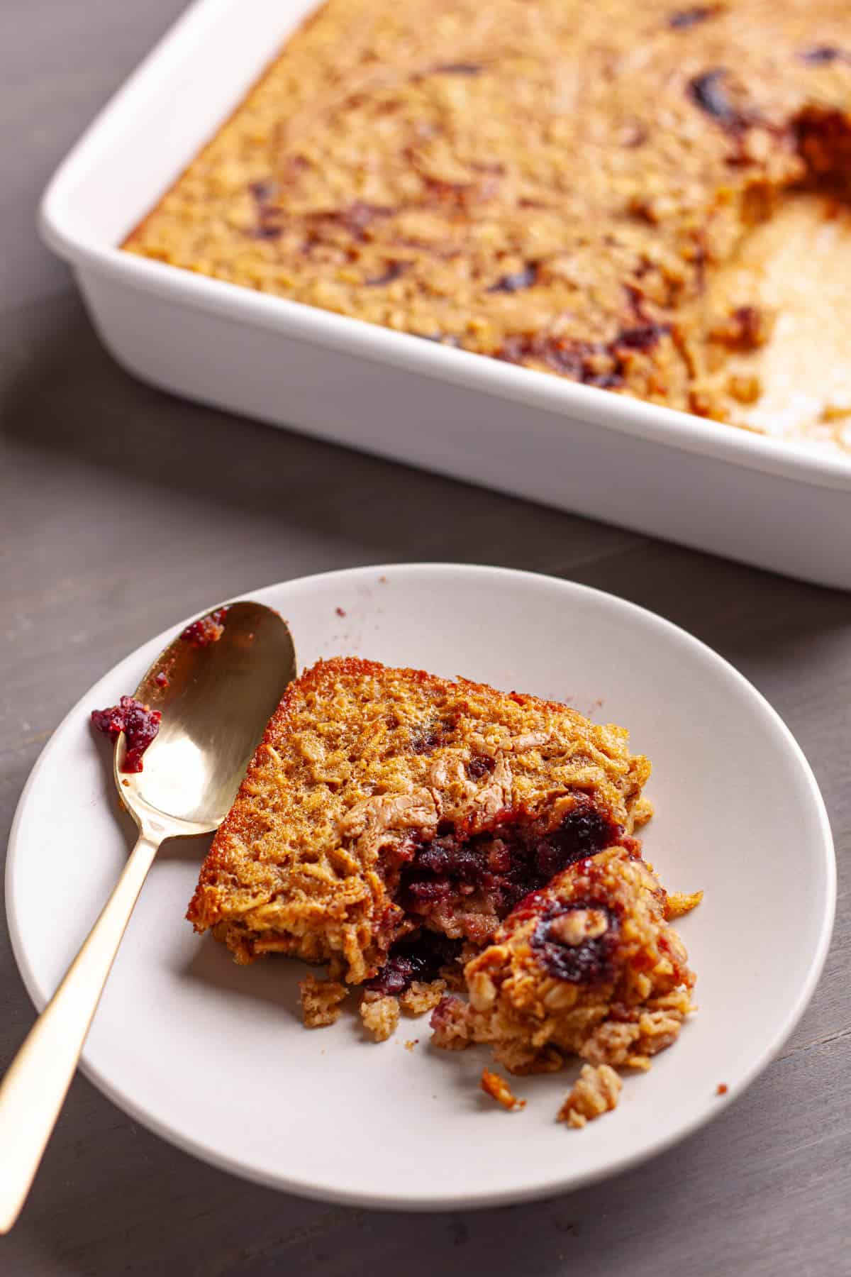 A piece of peanut butter and jam baked oatmeal on a small plate with a gold spoon.
