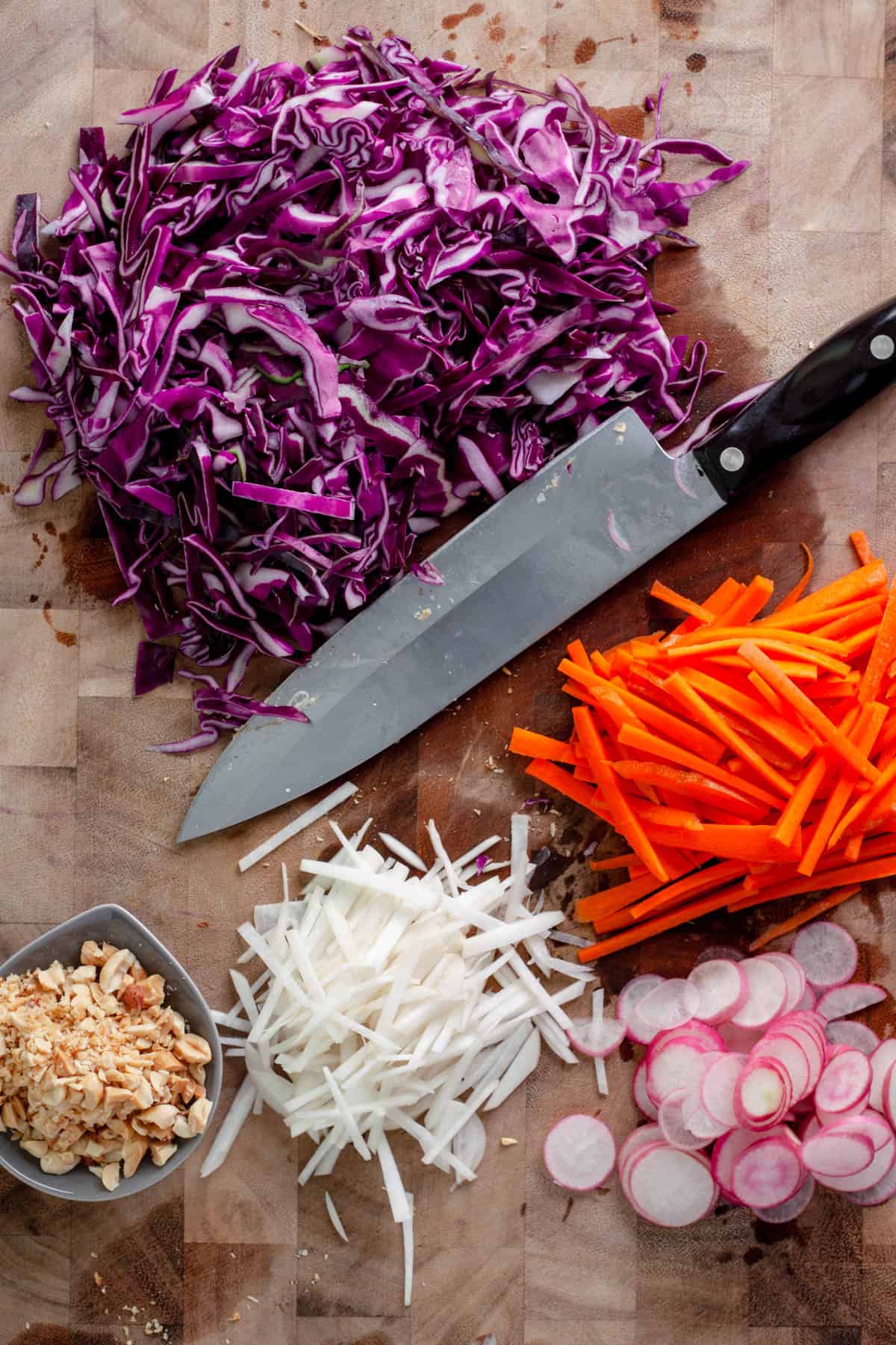 A cutting board and chef knife with sliced cabbage, carrots, turnips, radishes and peanuts.