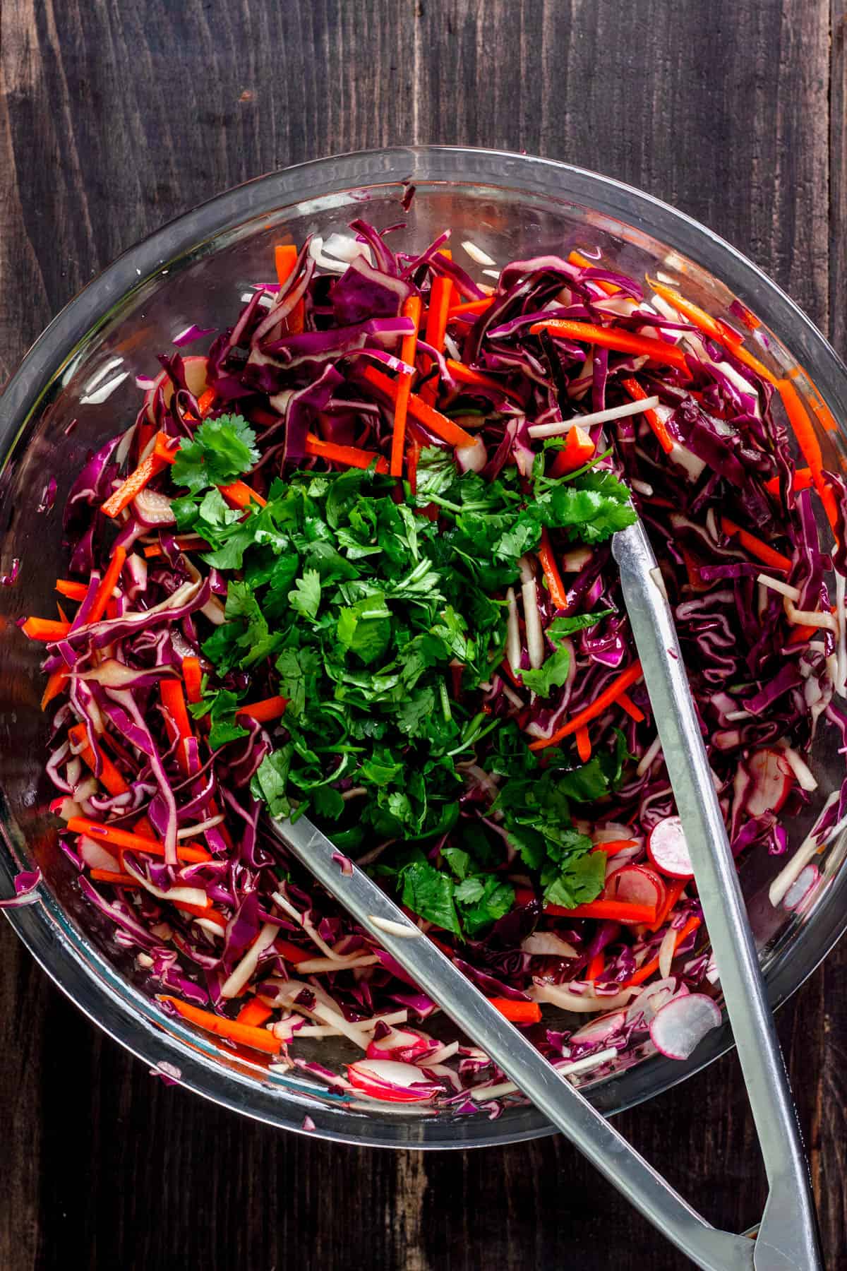 A purple cabbage slaw in a large mixing bowl with tongs getting ready to toss it together.