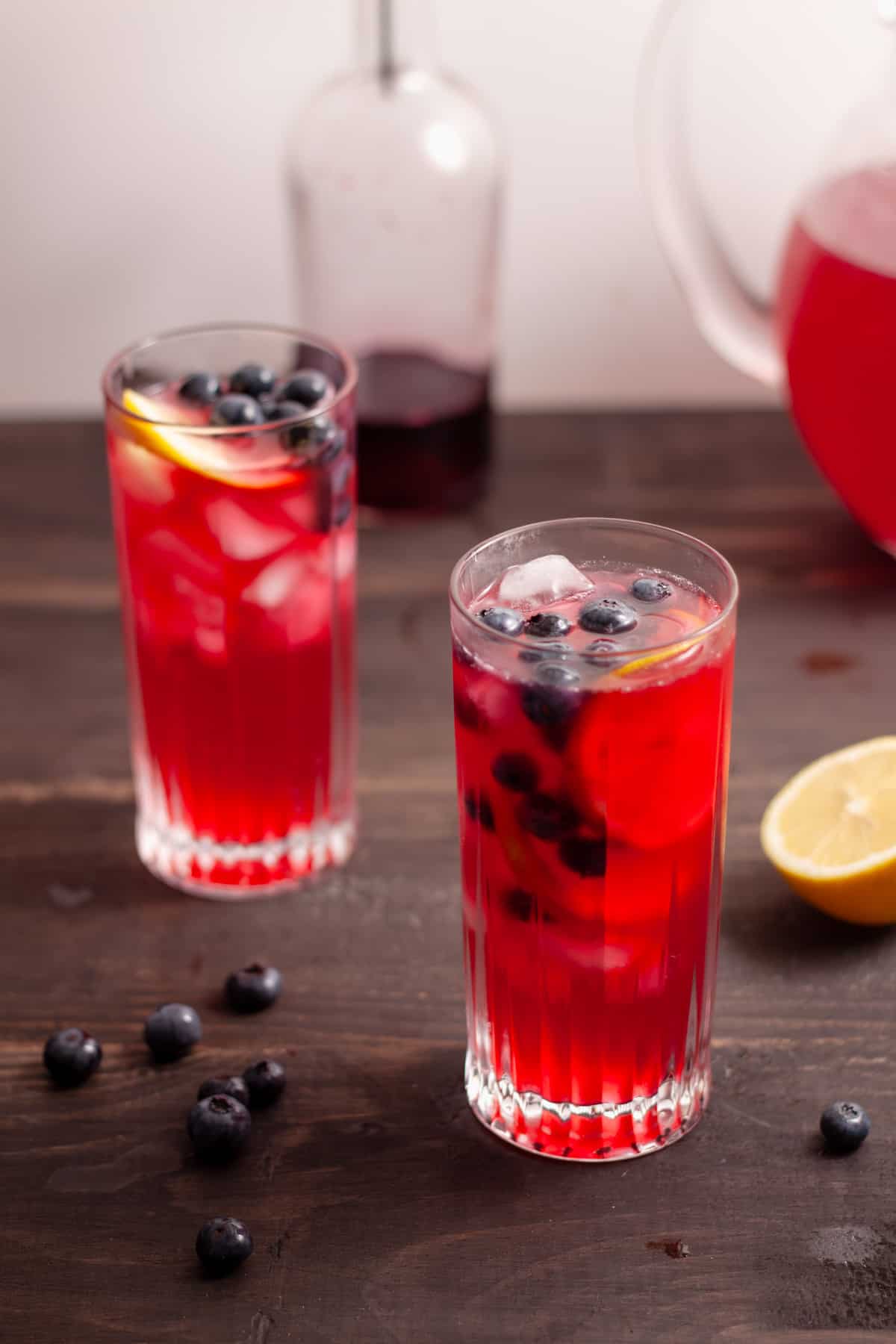 Homemade blueberry lemonade in a couple tall glasses and a bottle of blueberry syrup in the background.