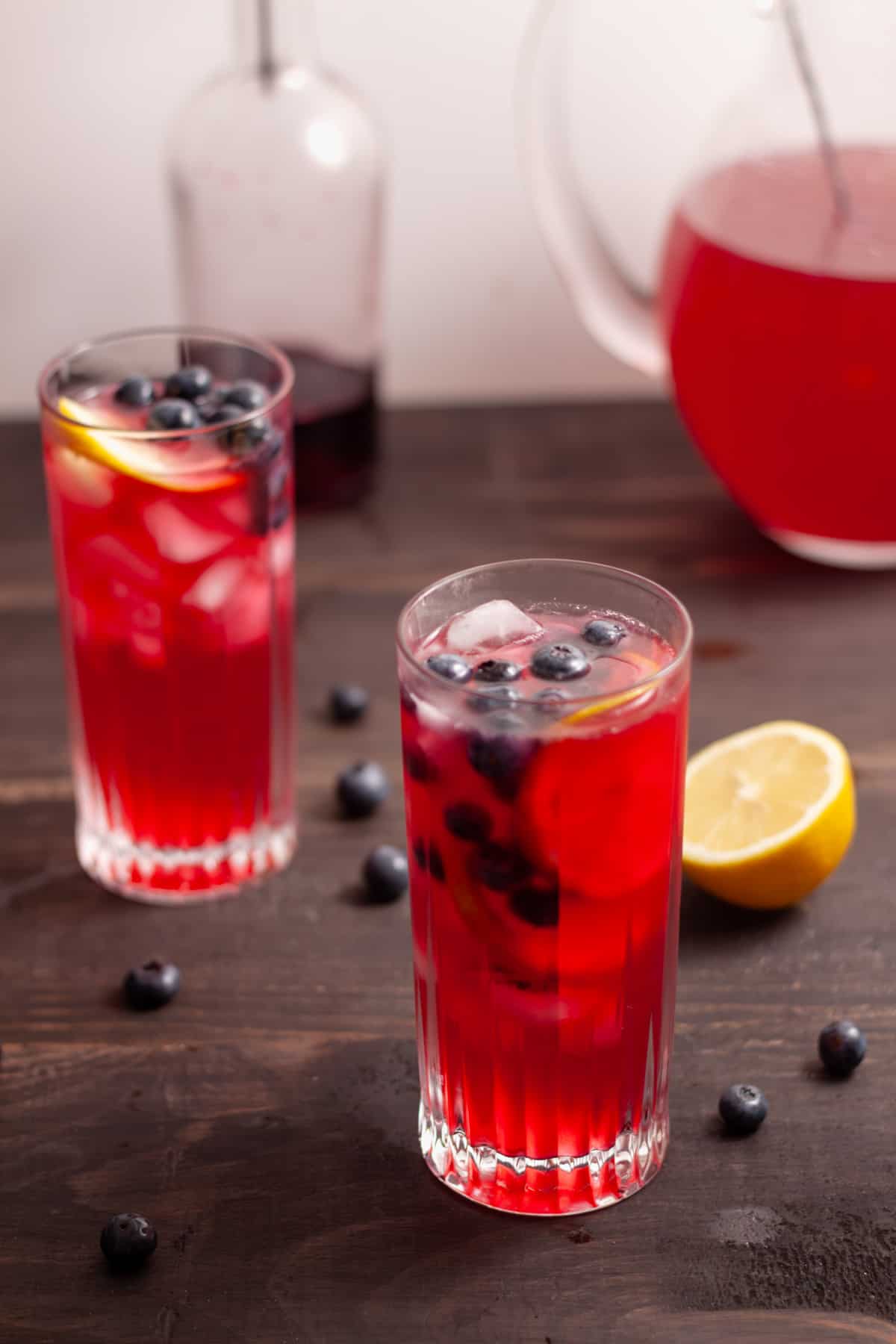 Homemade blueberry lemonade in a couple tall glasses and a bottle of blueberry syrup in the background.