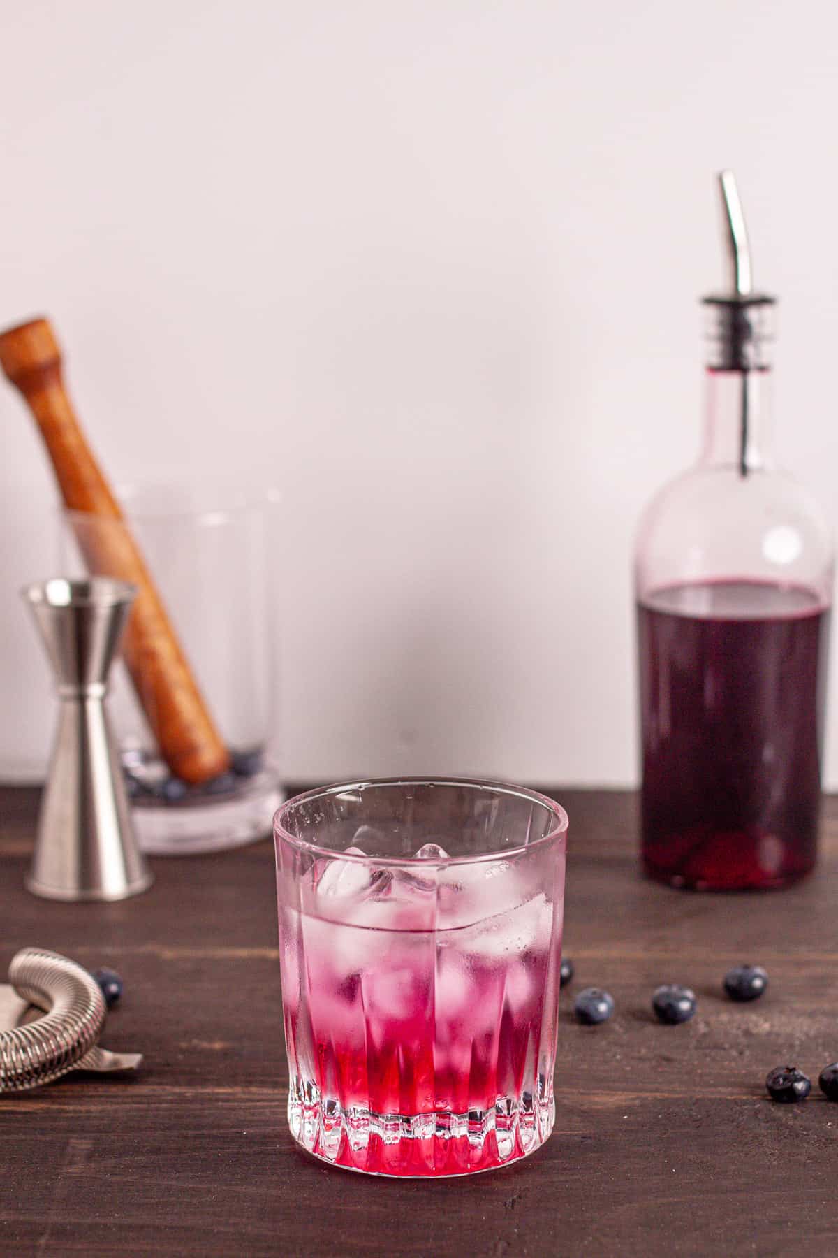 A glass of ice and water with blueberry simple syrup and a bottle of blueberry syrup in the background.