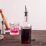 A bottle of blueberry simple syrup with a glass of blueberry lemonade in the background.