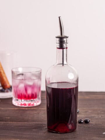 A bottle of blueberry simple syrup with a glass of blueberry lemonade in the background.