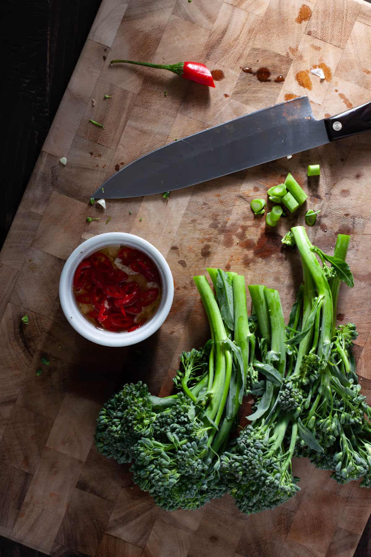 Trimmed broccolini on a cutting board with a small white bowl holding sliced chili peppers, garlic and oil.