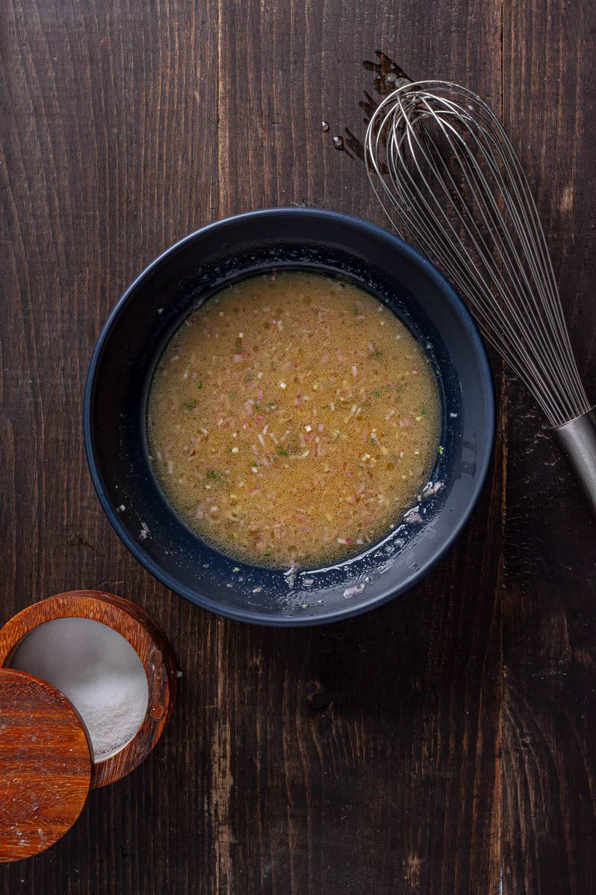A white balsamic vinaigrette in a medium mixing bowl with a dish of salt to the side and a whisk off the bowl.