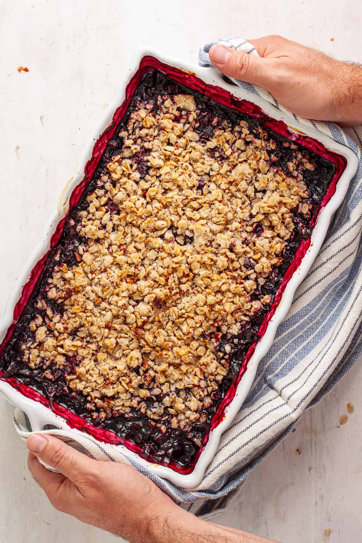 Two hands holding the edges of a white baking dish with a vegan blueberry crisp inside.