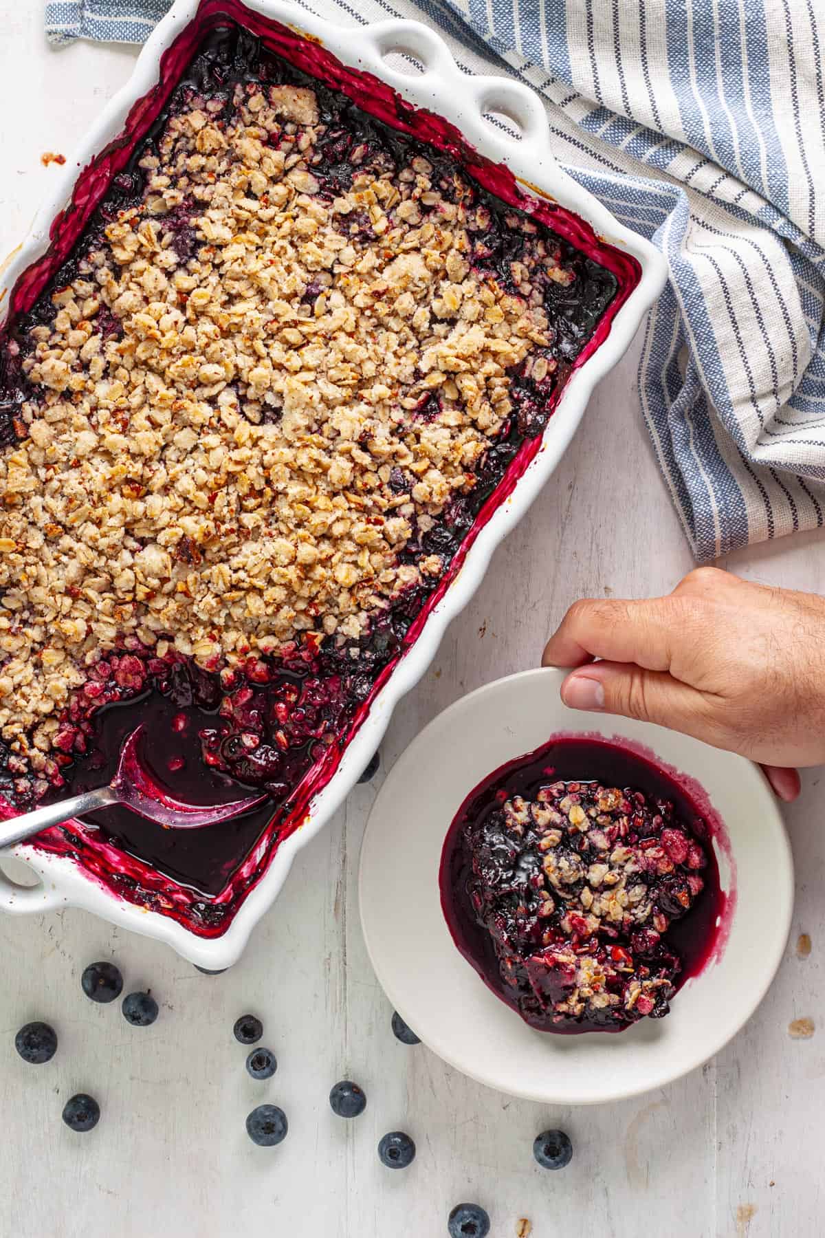 A hand setting down a small plate with a serving of vegan blueberry crisp and the full baking dish off to the side.