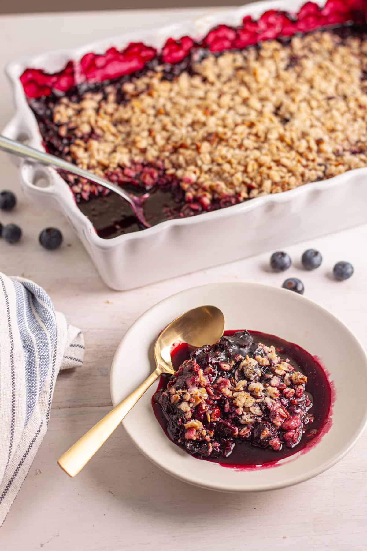 A serving of vegan blueberry crisp on a small plate with the full baking dish in the background.