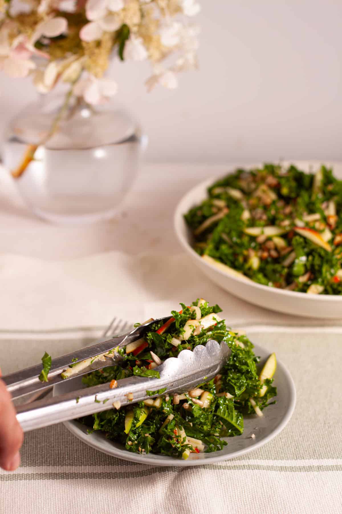 Tongs serving kale and apple slaw onto a small plate with a larger serving bowl in the background.