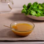 Maple Dijon Dressing in a small bowl on a table with a salad and flowers in the background.