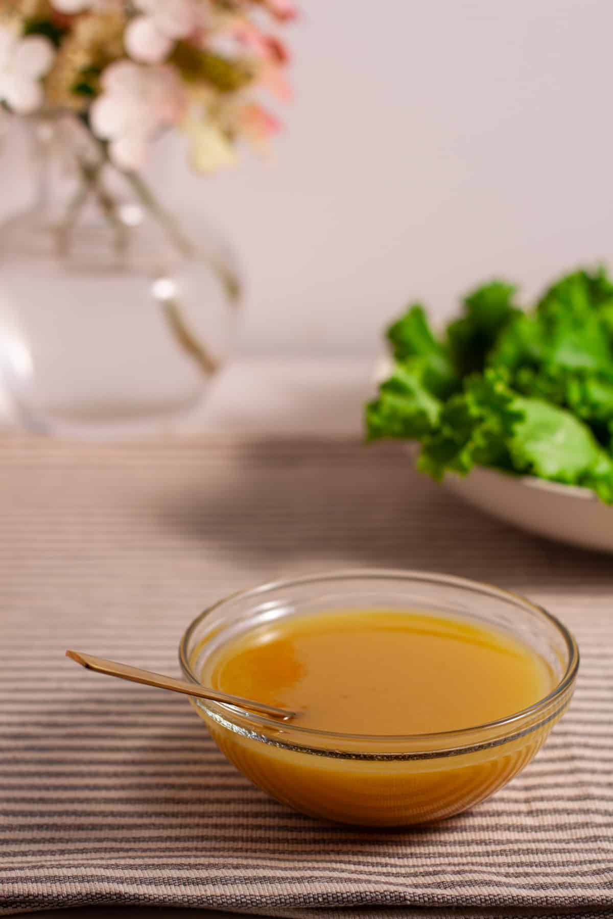 Maple Dijon Dressing in a small bowl on a table with a salad and flowers in the background.