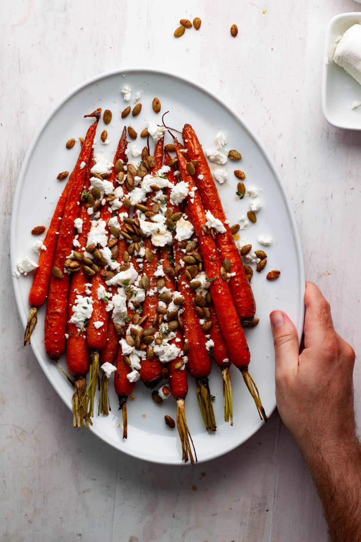 Maple roasted carrots on a white platter topped with goat cheese and pepitas with a hand pushing it onto a white table.
