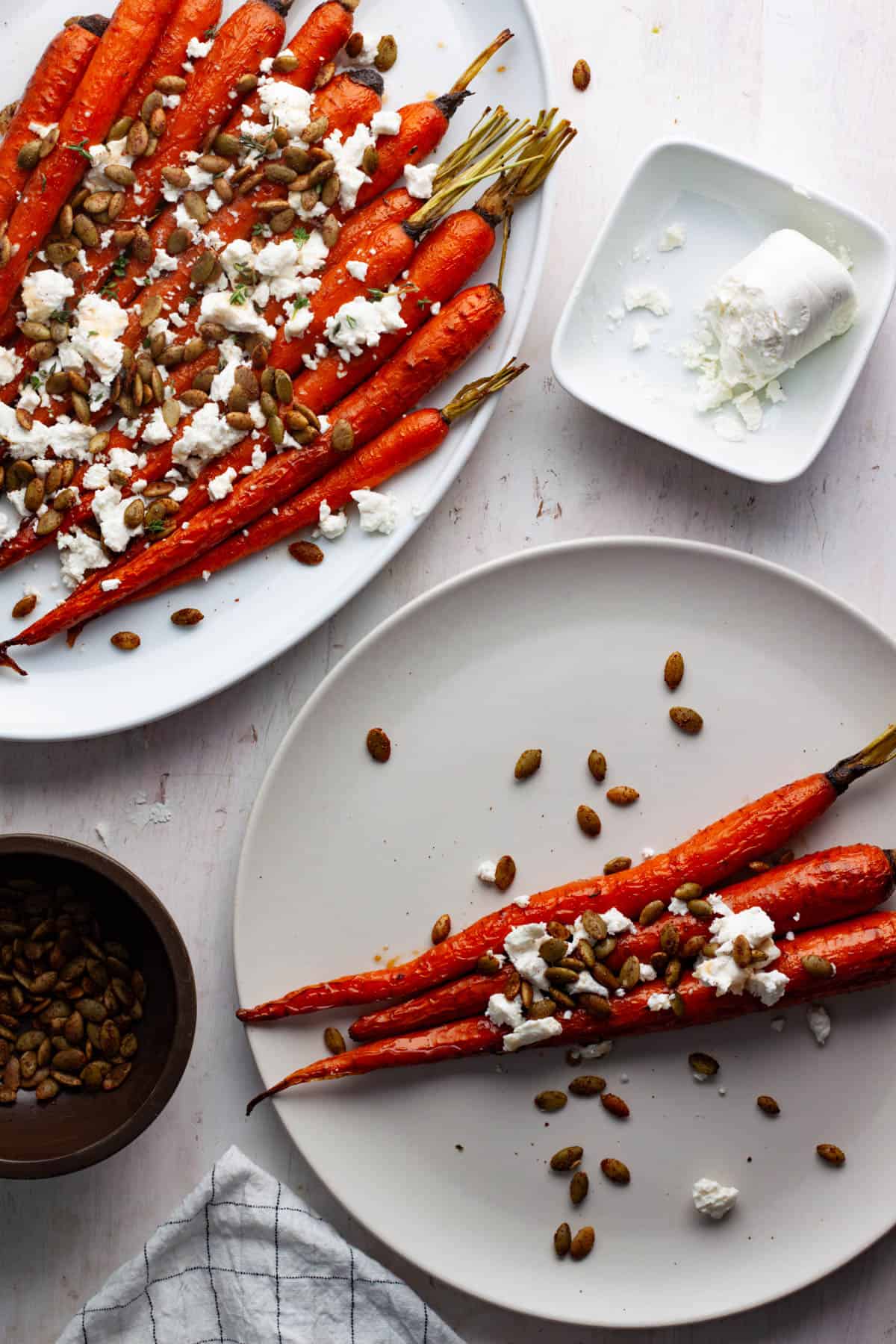 A serving of maple roasted carrots topped with crumbled goat cheese and pepitas with a platter of more carrots to the side.