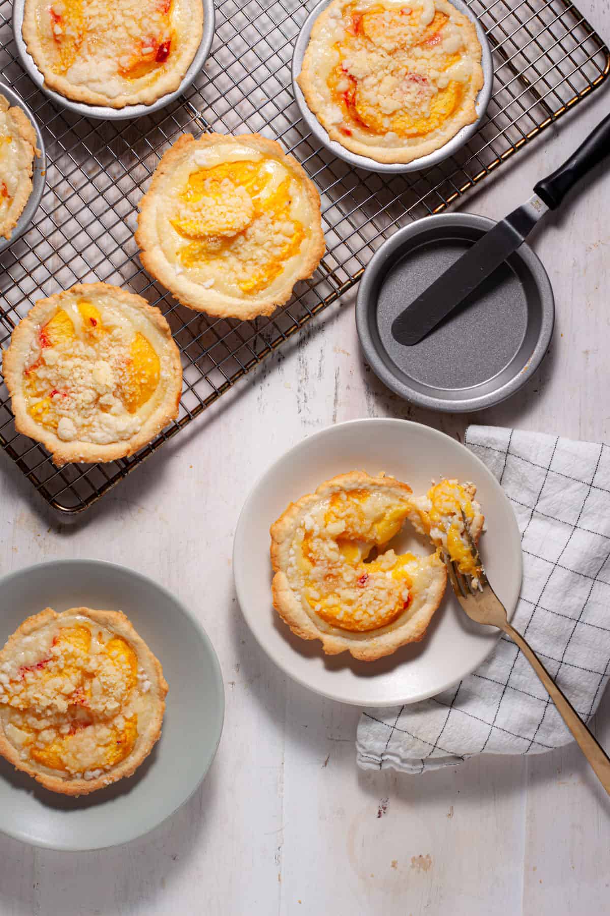 Mini peach custard pies cooling on a rack with a couple portions on plates and a fork cutting into one.