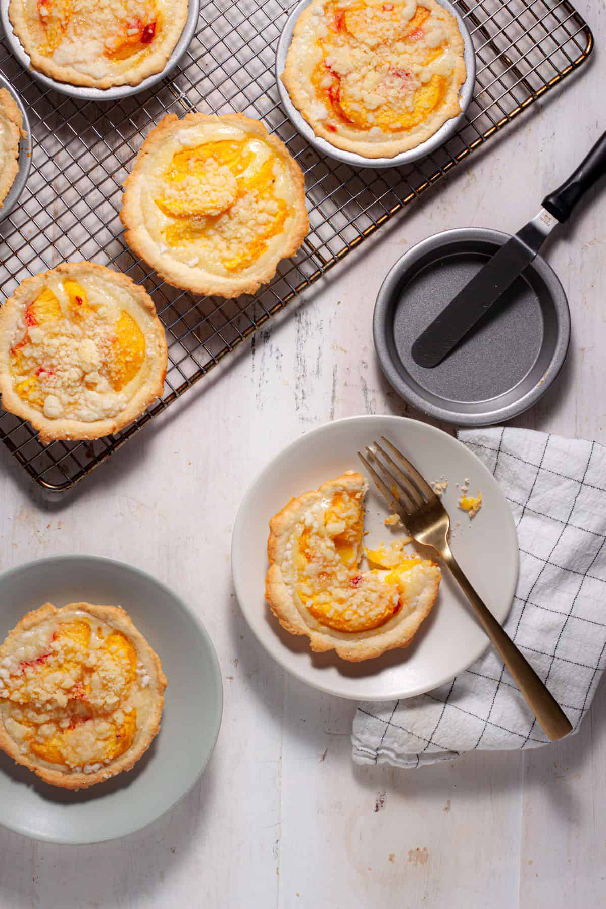Mini peach custard pies cooling on a rack with a couple portions on plates and a fork cutting into one.