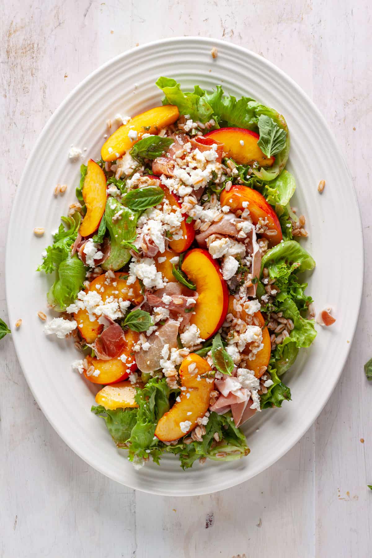 An assembled peach and goat cheese salad with prosciutto and farro on a white platter.