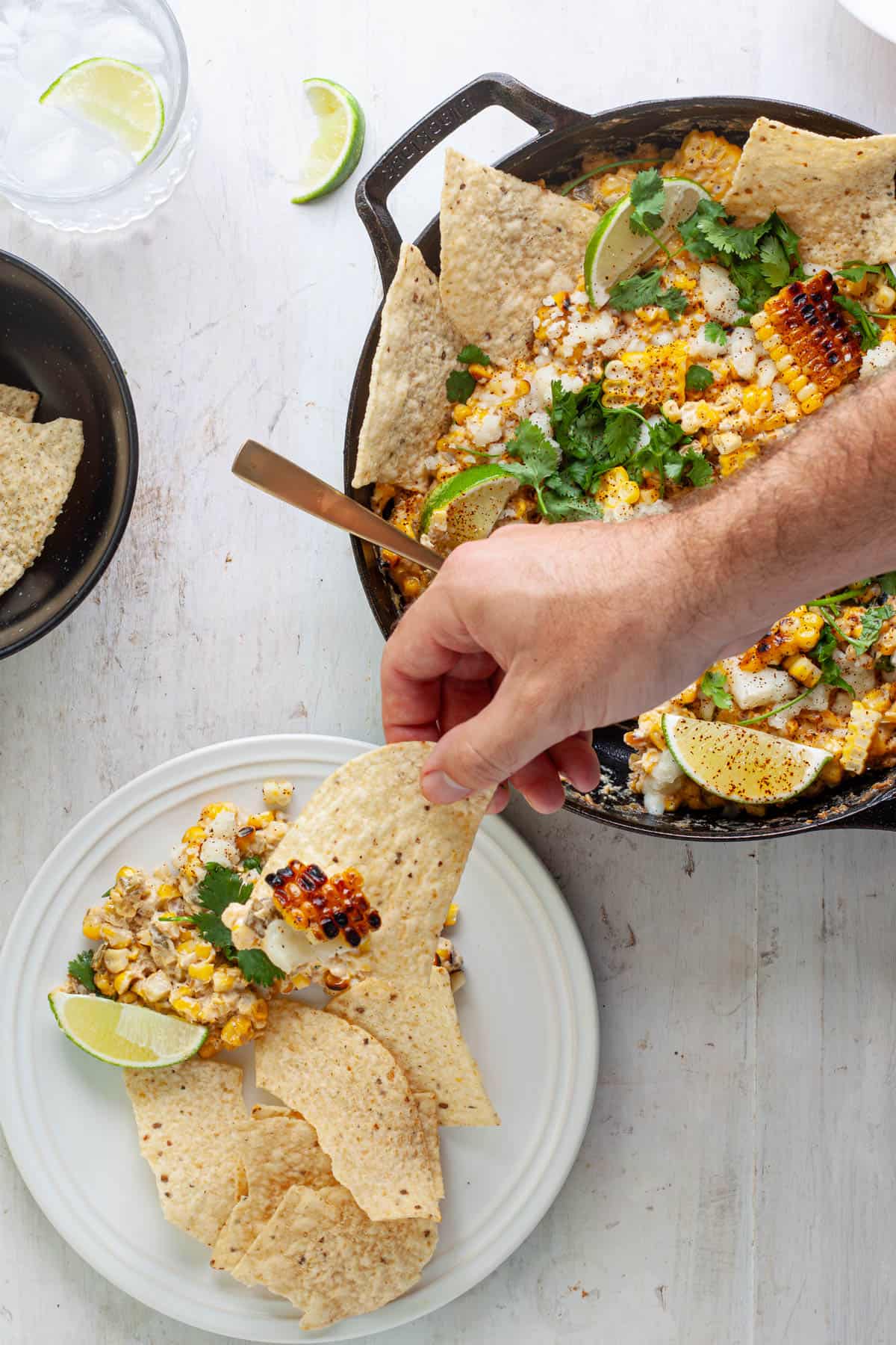 A hand grabbing a chip with street corn dip and a skillet with more dip to the side.