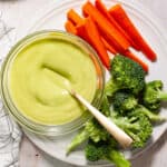 Avocado aioli getting stirred in a small bowl on a white plate with fresh veggies on the side.