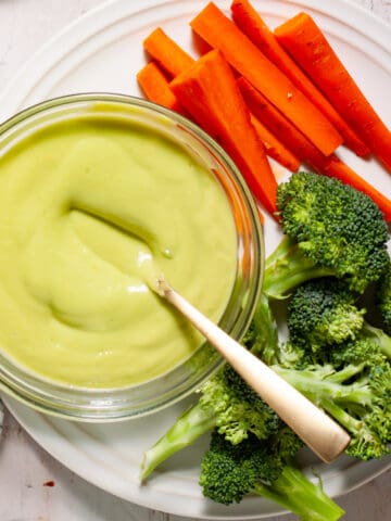 Avocado aioli getting stirred in a small bowl on a white plate with fresh veggies on the side.