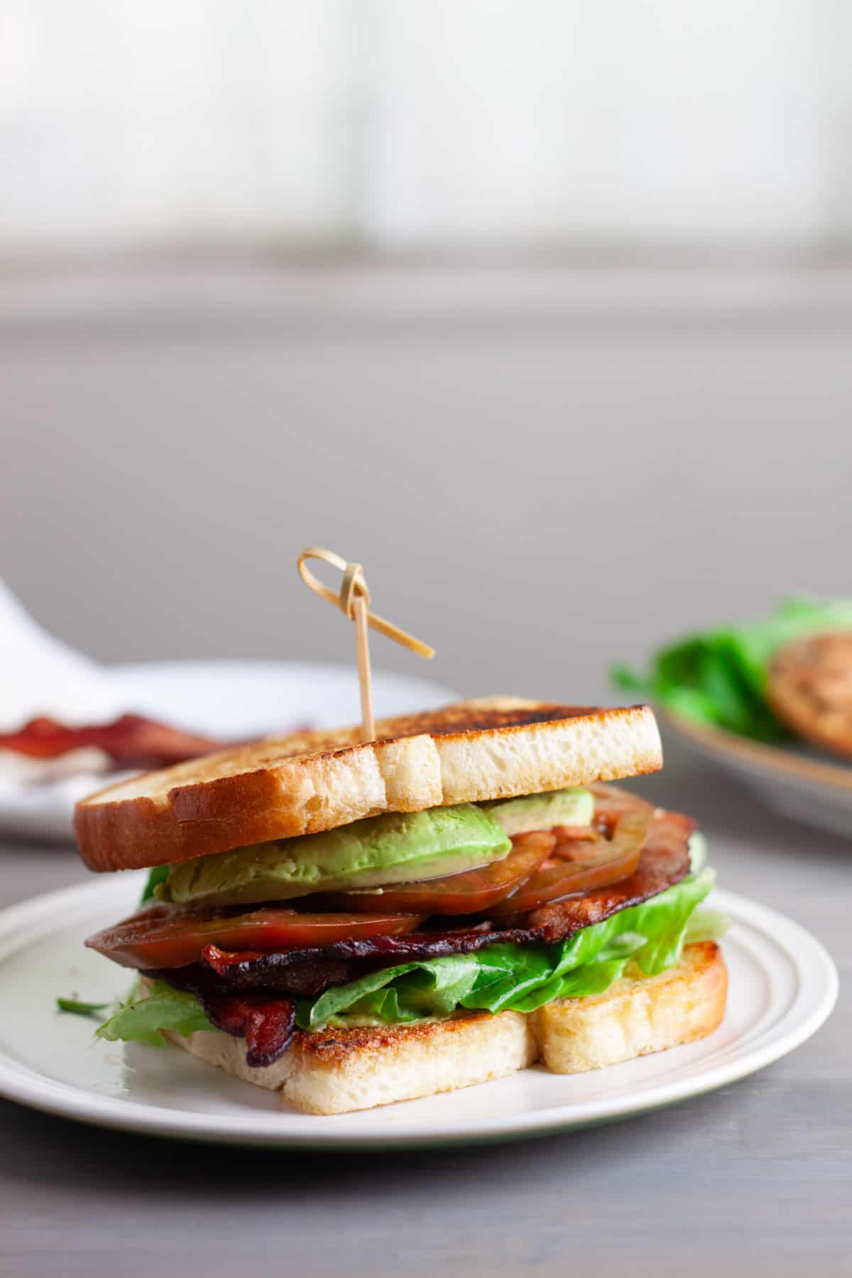 An avocado BLT on a toasted white bread on top of a white plate.
