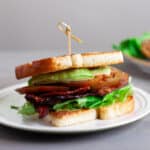 An avocado BLT on a toasted white bread on top of a white plate.