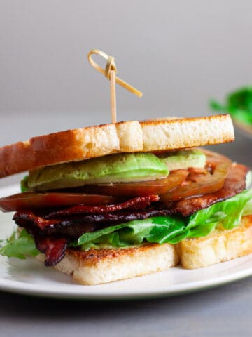 An avocado BLT on a toasted white bread on top of a white plate.